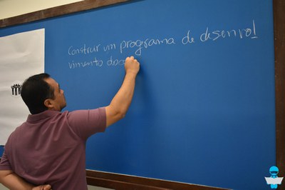 Foto de um Professor, com uma camiseta polo roxa, escrevendo na ousa azul "Construir um programa de desenvolvimento", com giz branco.
