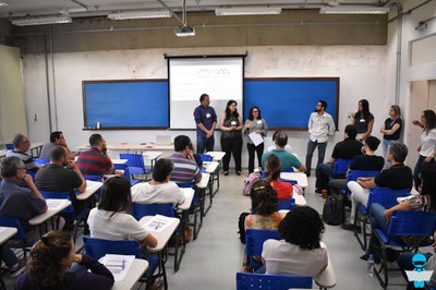 Foto de uma sala de aula, com vários professores sentados em carteiras escolares, e 7 professores em pé, na frente da lousa azul.