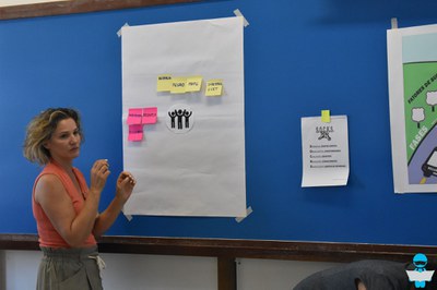 Foto de uma Professora, explicando os cartazes com post its coloridos, colados na lousa azul.