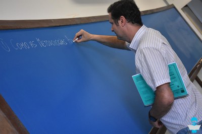 Foto de um Professor, com uma camisa branca listrada, escrevendo na lousa azul, com giz branco.