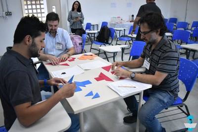 Foto de 3 professores em uma mesa, manuseando papeis coloridos recortados em formas geométricas.