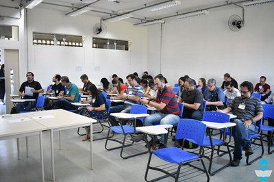 Foto dos professores em uma sala de aula, sentados em carteiras escolares azuis.