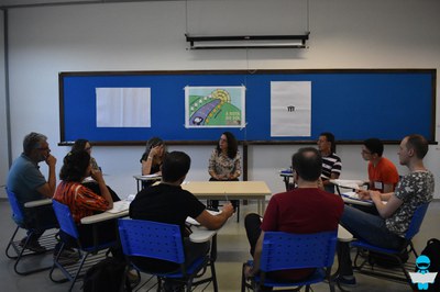 Foto de uma roda de professores, sentados em carteiras escolares. Ao fundo uma lousa azul, com cartazes colados.