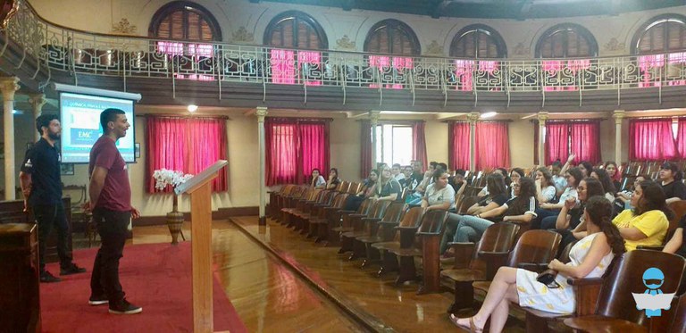 Foto da palestra ocorrendo em um auditório. O auditório possui piso e cadeiras fixas de madeira, dois andares contendo ao fundo 12 janelas com cortinas vermelhas. À esquerda dois palestrantes sobre um tapete vermelho, em frente à uma tela de projeção, um homem veste camiseta bordô do curso Engenharia de Materiais e calça preta, o outro veste uma camiseta polo azul marinho e calça preta. À direita plateia atenta sentada nas cadeiras.