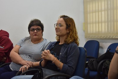 Foto de duas mulheres durante a roda de conversa. À esquerda uma mulher de cabelo curto, usando óculos e vestindo uma camiseta listrada. Ao seu lado uma mulher com cabelos claros, óculos, vestindo uma camiseta preta e uma camisa xadrez azul, suas mãos estão juntas na frente do peito.